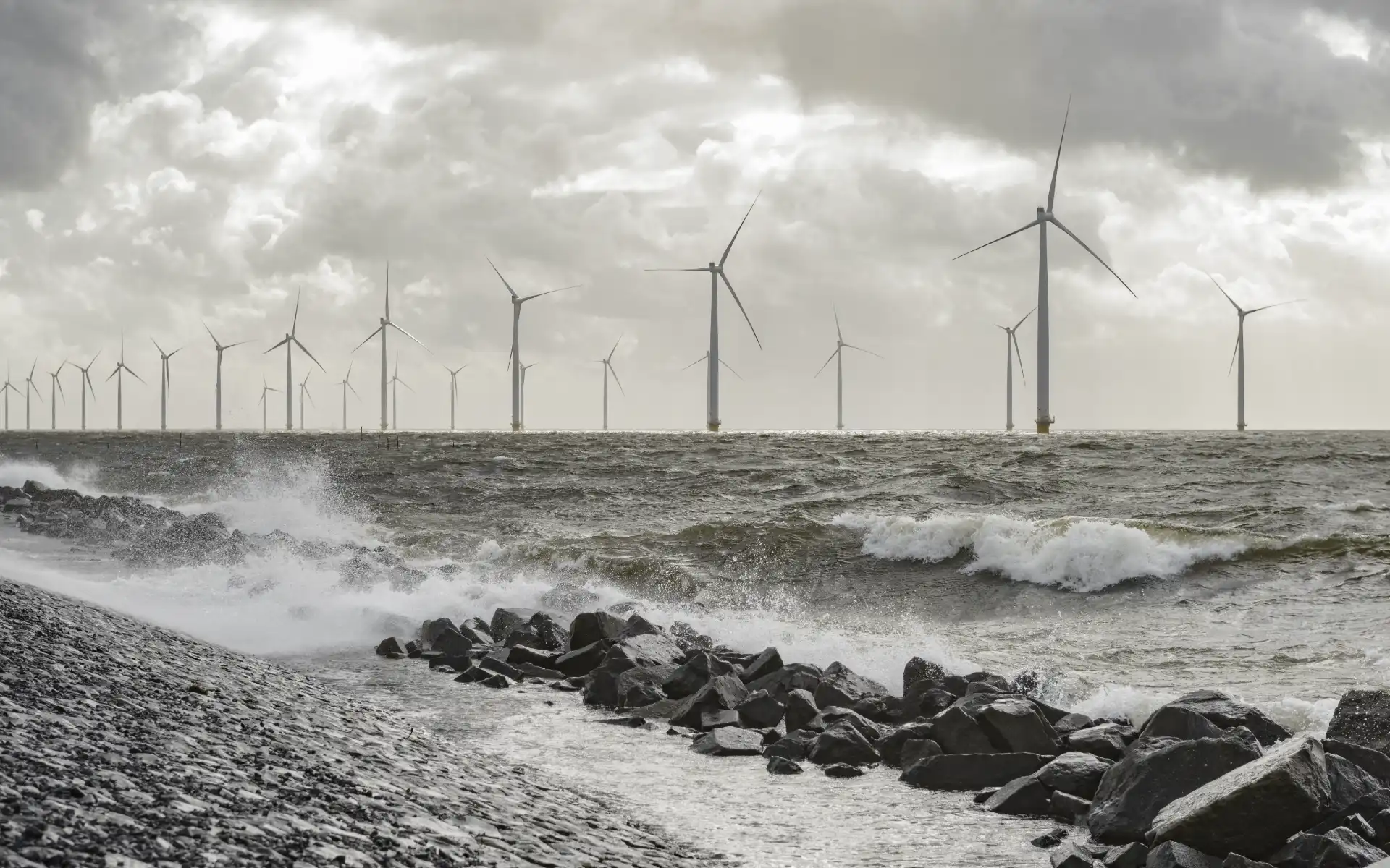 Offshore wind farm with turbines standing in rough sea conditions, waves crashing against rocky shoreline under dramatic clouds.