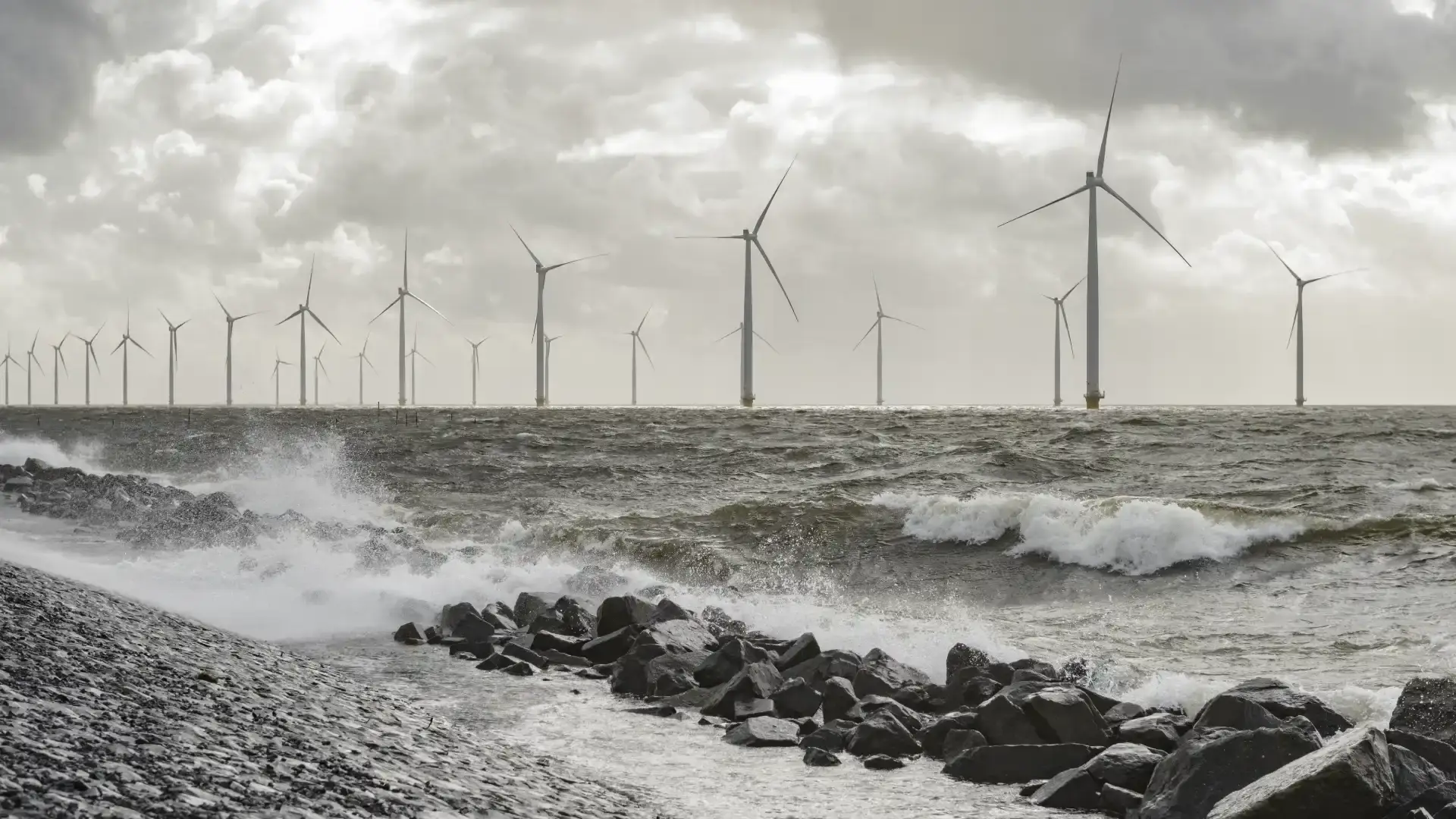 Offshore wind farm with turbines standing in rough sea conditions, waves crashing against rocky shoreline under dramatic clouds.