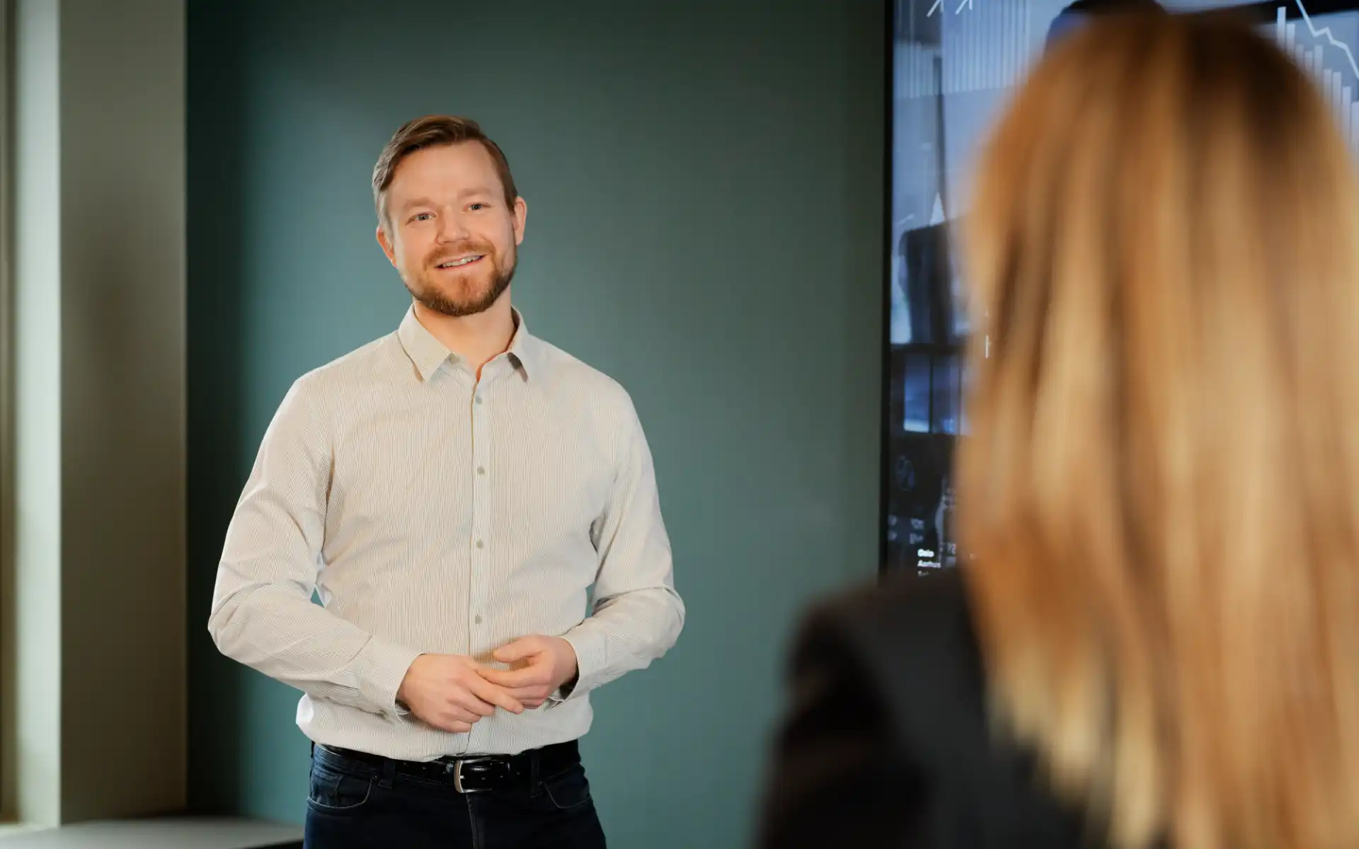 Man standing and speaking in a meeting room with a presentation screen in the background.
