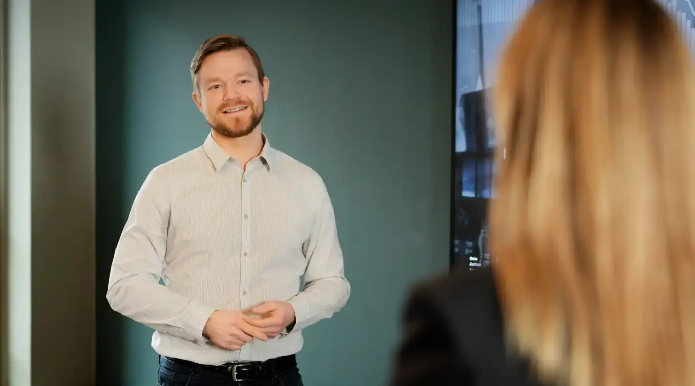 Man standing and speaking in a meeting room with a presentation screen in the background.