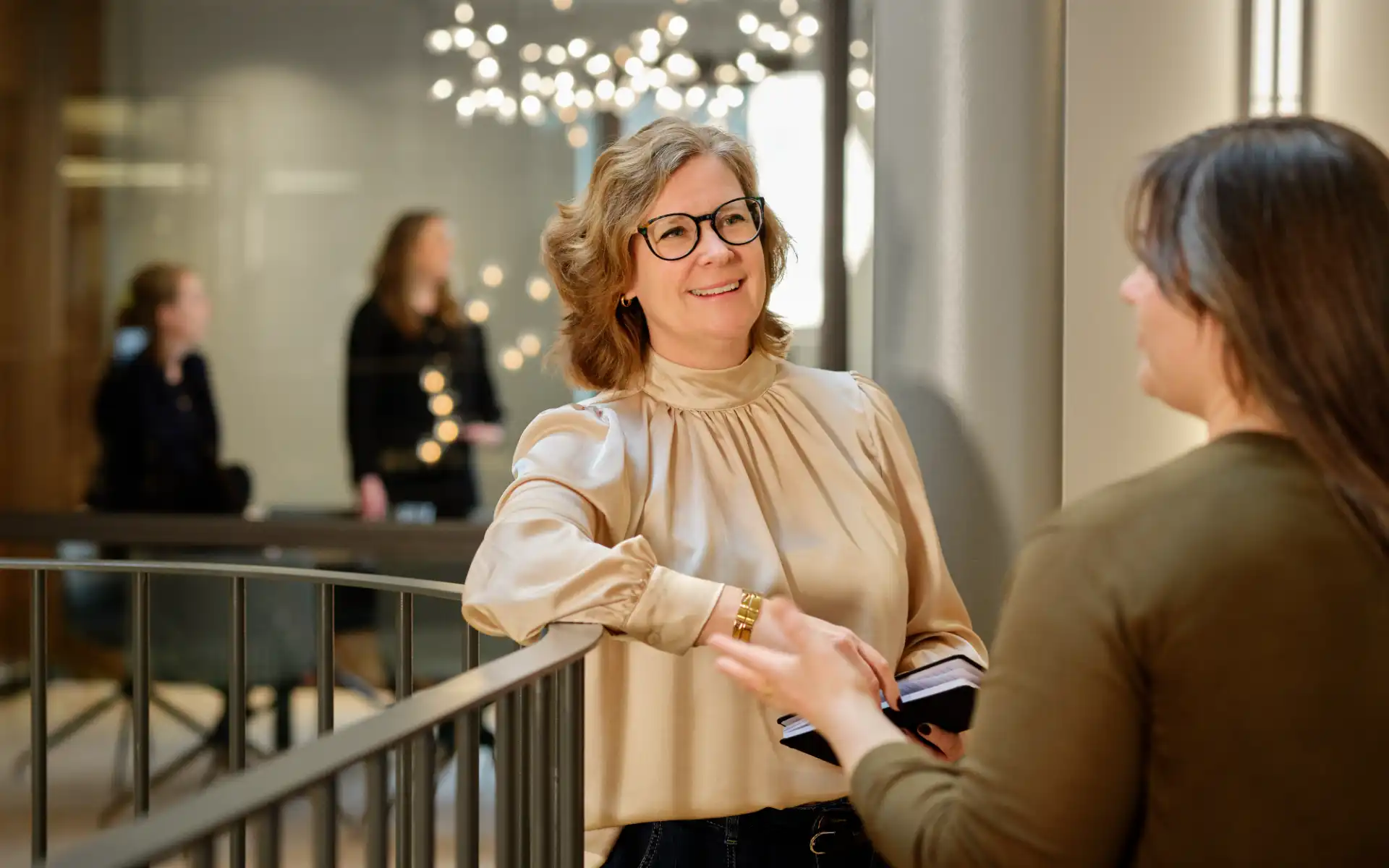 Two women talking and smiling by a railing in a bright office environment.
