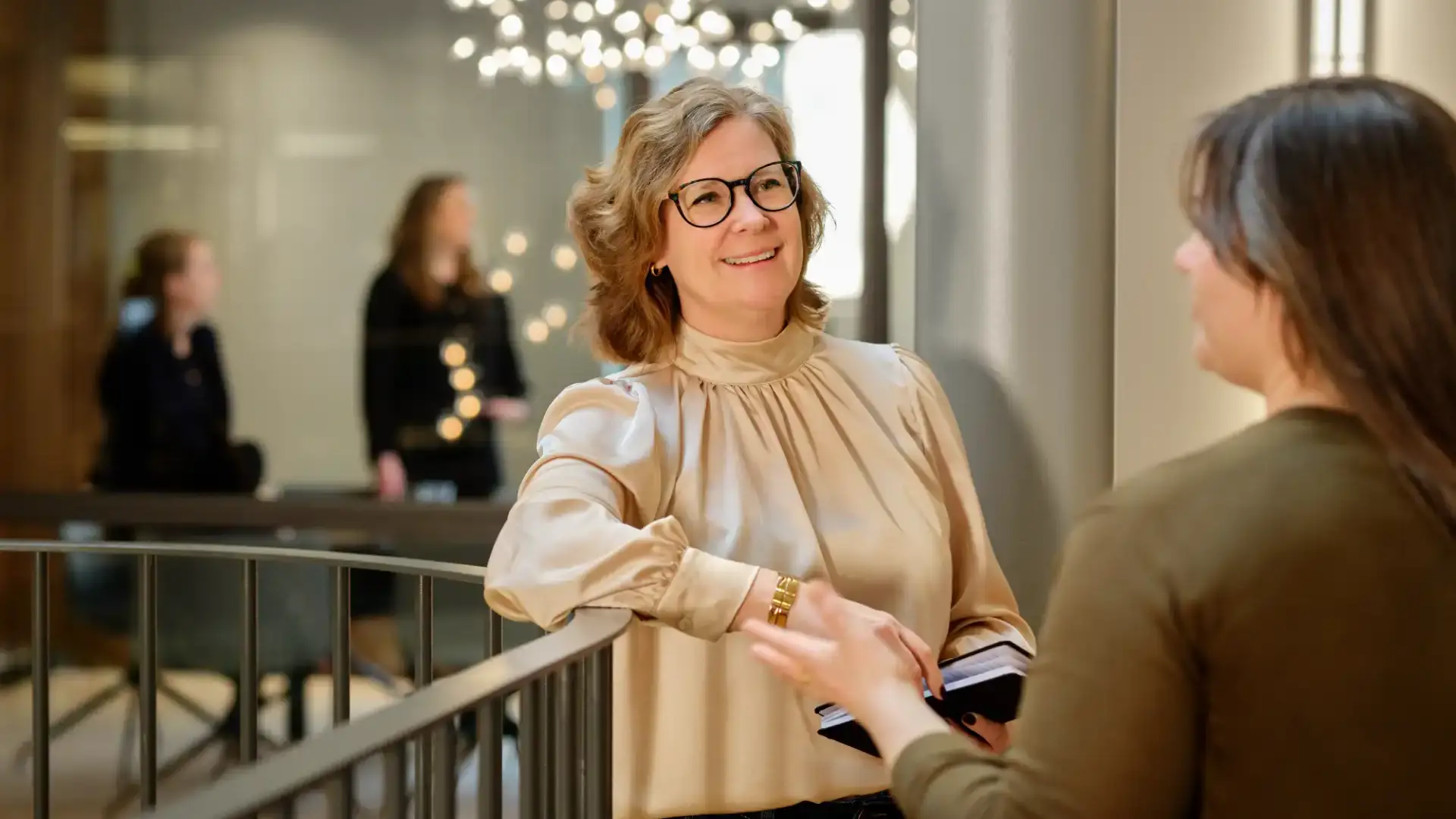 Two women talking and smiling by a railing in a bright office environment.