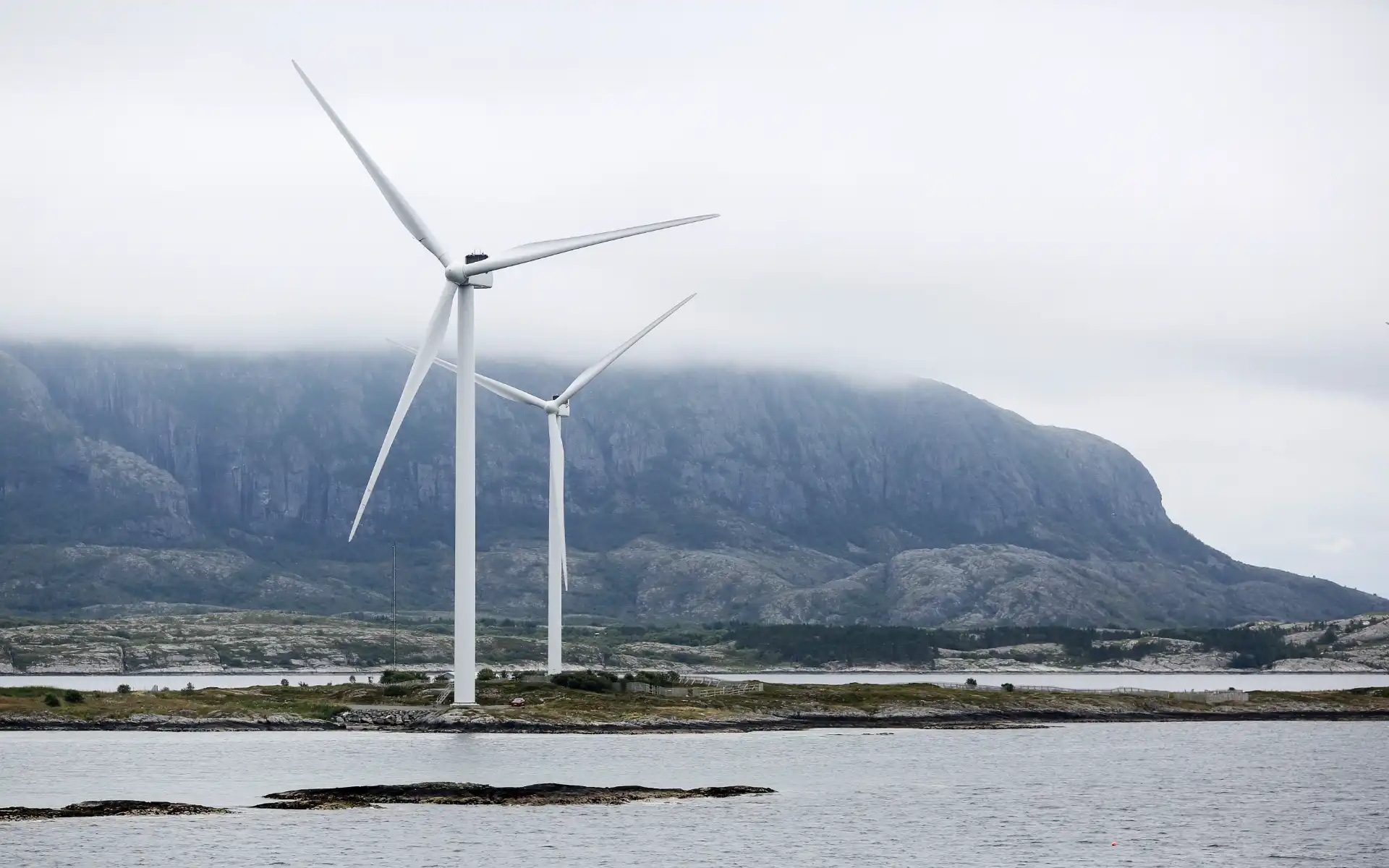 Offshore wind turbines rising above the coastline with misty mountains in the background.