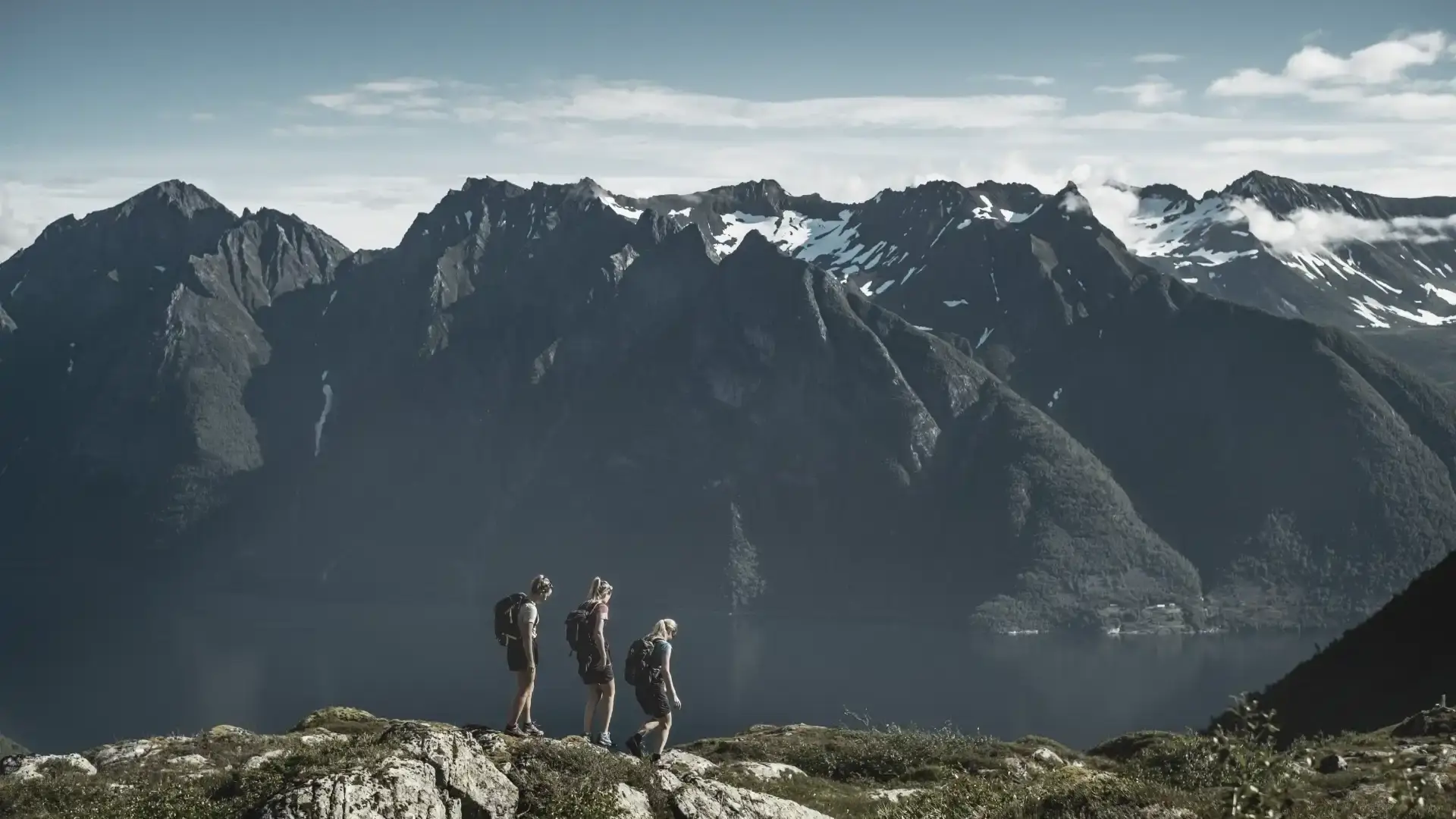 Three hikers walking along a ridge with dramatic mountain peaks and a fjord below, reflecting unity, resilience, and the power of nature in renewable energy.