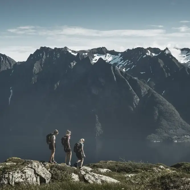 Three hikers walking along a ridge with dramatic mountain peaks and a fjord below, reflecting unity, resilience, and the power of nature in renewable energy.
