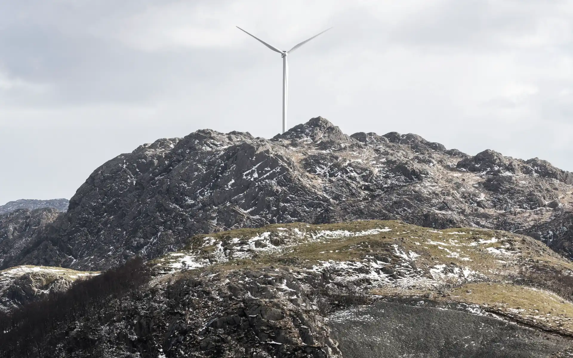 Wind turbine standing on a rugged, snow-dusted mountain peak under an overcast sky.