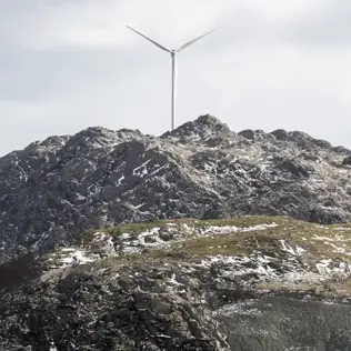 Wind turbine standing on a rugged, snow-dusted mountain peak under an overcast sky.