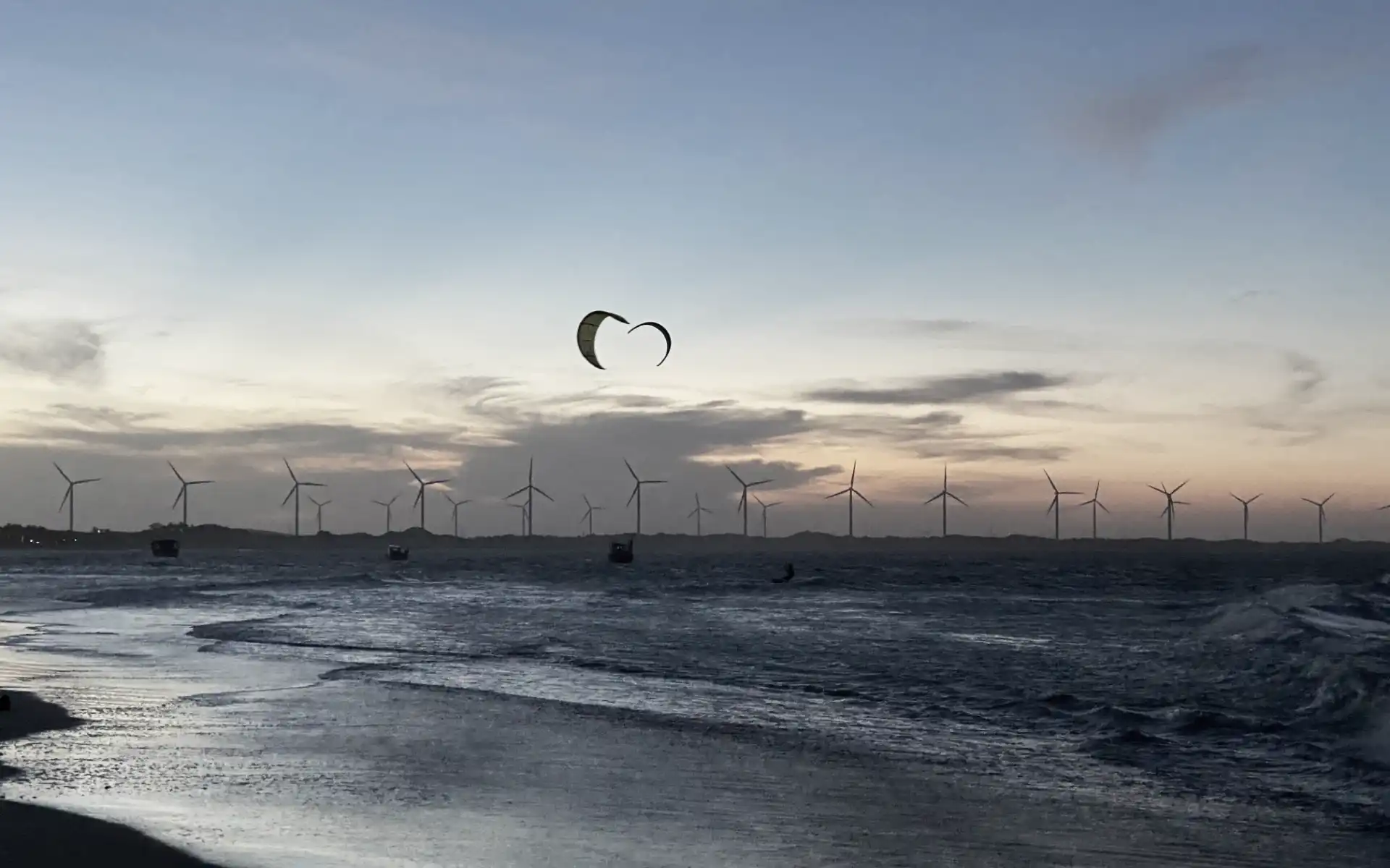 Silhouetted offshore wind turbines at sunset with kite surfers gliding across the water near the shoreline.