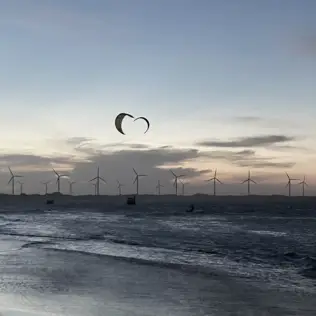 Silhouetted offshore wind turbines at sunset with kite surfers gliding across the water near the shoreline.