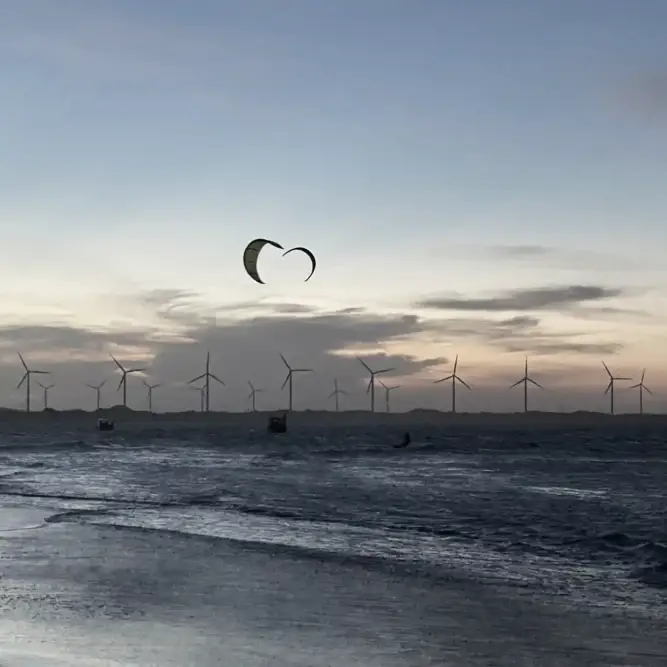 Silhouetted offshore wind turbines at sunset with kite surfers gliding across the water near the shoreline.